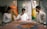 A group of three people is gathered around a table, focusing intently on an art piece or textile in a serene setting. Two mannequins dressed in traditional, colorful clothing are positioned in the background. The atmosphere suggests a blend of traditional craft and modern presentation.