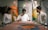 A group of three people is gathered around a table, focusing intently on an art piece or textile in a serene setting. Two mannequins dressed in traditional, colorful clothing are positioned in the background. The atmosphere suggests a blend of traditional craft and modern presentation.