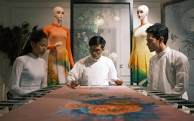 A group of three people is gathered around a table, focusing intently on an art piece or textile in a serene setting. Two mannequins dressed in traditional, colorful clothing are positioned in the background. The atmosphere suggests a blend of traditional craft and modern presentation.