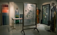 A dimly lit room features mannequins dressed in artistic clothing, showcasing a collection of garments with intricate patterns and vibrant colors. A decorative frame with frosted glass and bird and plant motifs stands in the center, surrounded by vibrant textiles and art pieces. The setting exudes an elegant and sophisticated atmosphere.