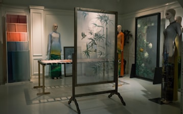 A dimly lit room features mannequins dressed in artistic clothing, showcasing a collection of garments with intricate patterns and vibrant colors. A decorative frame with frosted glass and bird and plant motifs stands in the center, surrounded by vibrant textiles and art pieces. The setting exudes an elegant and sophisticated atmosphere.