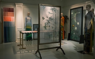A dimly lit room features mannequins dressed in artistic clothing, showcasing a collection of garments with intricate patterns and vibrant colors. A decorative frame with frosted glass and bird and plant motifs stands in the center, surrounded by vibrant textiles and art pieces. The setting exudes an elegant and sophisticated atmosphere.