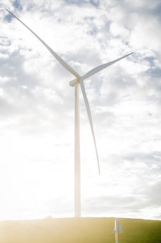 A large wind turbine stands prominently against a partly cloudy sky. The structure towers over a grassy hill, highlighted by bright sunlight reflecting off its white blades.