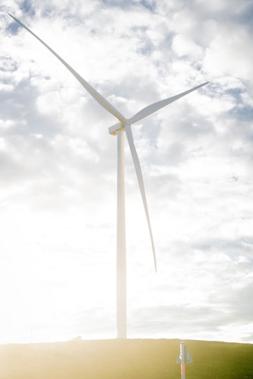 A large wind turbine stands prominently against a partly cloudy sky. The structure towers over a grassy hill, highlighted by bright sunlight reflecting off its white blades.