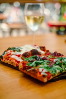 A close-up view of a rectangular pizza topped with arugula, tomatoes, cheese, and other ingredients, placed on a wooden surface. In the background, there is a blurry glass of white wine, suggesting a casual dining setting.