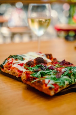 A close-up view of a rectangular pizza topped with arugula, tomatoes, cheese, and other ingredients, placed on a wooden surface. In the background, there is a blurry glass of white wine, suggesting a casual dining setting.