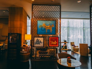 A cozy and stylish indoor setting features modern artwork on a decorative partition. The artwork includes a blue framed piece depicting an abstract animal, a red framed piece with floral and animal motifs, and a landscape scene. The decor includes a barrel with a lamp and wine bottles, luxurious seating, and ambient lighting from large windows with sheer curtains.