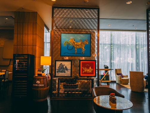 A cozy and stylish indoor setting features modern artwork on a decorative partition. The artwork includes a blue framed piece depicting an abstract animal, a red framed piece with floral and animal motifs, and a landscape scene. The decor includes a barrel with a lamp and wine bottles, luxurious seating, and ambient lighting from large windows with sheer curtains.