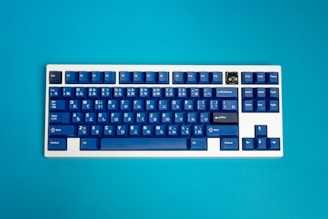A mechanical keyboard with blue keycaps featuring Japanese hiragana characters is set against a vibrant turquoise background. The keyboard includes a distinct key with a unique design in the top right corner and a traditional layout with function keys at the top.