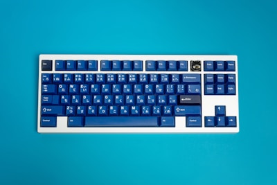 A mechanical keyboard with blue keycaps featuring Japanese hiragana characters is set against a vibrant turquoise background. The keyboard includes a distinct key with a unique design in the top right corner and a traditional layout with function keys at the top.