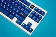 A mechanical keyboard featuring blue keycaps with a mix of English and Japanese characters is positioned against a bright blue background. One keycap in the shape of a black cat with yellow eyes stands out among the others.