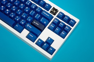 A mechanical keyboard featuring blue keycaps with a mix of English and Japanese characters is positioned against a bright blue background. One keycap in the shape of a black cat with yellow eyes stands out among the others.