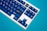 A mechanical keyboard featuring blue keycaps with a mix of English and Japanese characters is positioned against a bright blue background. One keycap in the shape of a black cat with yellow eyes stands out among the others.
