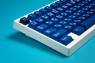 A sleek mechanical keyboard featuring deep blue keycaps with white Japanese characters and symbols on them. The keyboard has a modern, minimalist design with a white frame. It is set against a bright blue background, creating a clean and vibrant aesthetic.