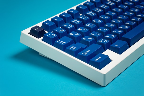 A sleek mechanical keyboard featuring deep blue keycaps with white Japanese characters and symbols on them. The keyboard has a modern, minimalist design with a white frame. It is set against a bright blue background, creating a clean and vibrant aesthetic.