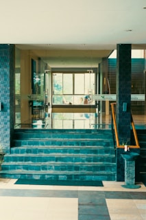A modern entrance area with a flight of tiled steps leading up to glass doors. Beyond the doors, there is a well-lit interior space with natural light coming from large windows. The foreground features a dark mat and blue-gray tiles adorning the steps and walls.