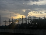 A silhouetted view of an electrical substation against a dramatic sky with the sun setting or rising, casting a warm glow behind the clouds.
