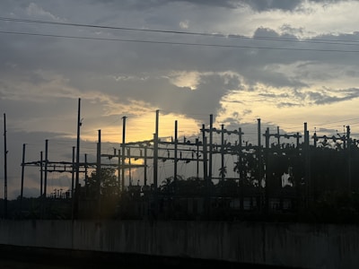 A silhouetted view of an electrical substation against a dramatic sky with the sun setting or rising, casting a warm glow behind the clouds.
