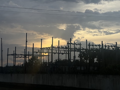 A silhouetted view of an electrical substation against a dramatic sky with the sun setting or rising, casting a warm glow behind the clouds.