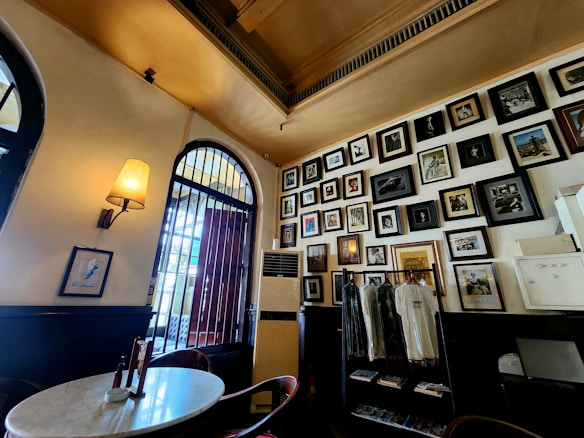 A cozy interior setting with a wall filled with an array of framed photographs, paintings, and artworks. A round marble table is set with condiments in the foreground, and there is a vintage-style lamp on the wall. Next to the door is a rack with hanging t-shirts.