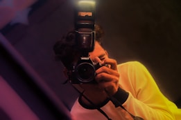 A person holds a camera with a flash attachment, capturing a photograph. The individual is wearing a light-colored shirt, and the image is composed with a slight tilt, featuring warm lighting and soft focus effects.