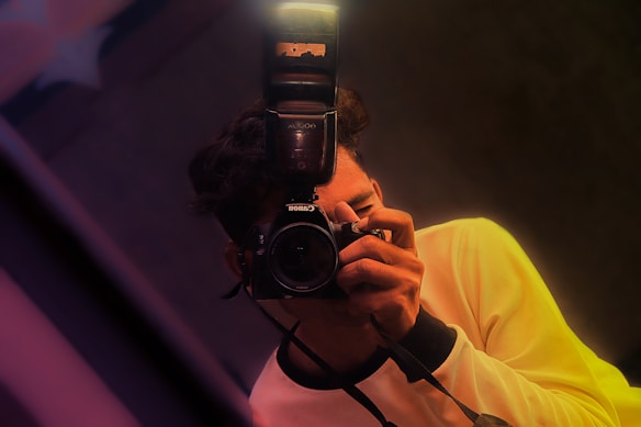 A person holds a camera with a flash attachment, capturing a photograph. The individual is wearing a light-colored shirt, and the image is composed with a slight tilt, featuring warm lighting and soft focus effects.