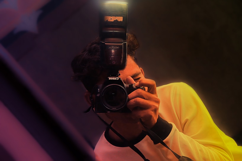 A person holds a camera with a flash attachment, capturing a photograph. The individual is wearing a light-colored shirt, and the image is composed with a slight tilt, featuring warm lighting and soft focus effects.
