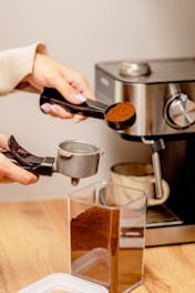 Two hands are preparing to make coffee by using a portafilter to scoop ground coffee from a container next to an espresso machine. The scene is set on a wooden countertop, and there's a mug placed under the spout of the machine.