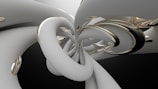 A digital abstract artwork featuring intertwined and looping shapes in metallic and off-white colors. The forms are smooth and glossy, creating a sense of depth and movement against a dark background.