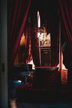A dimly lit, opulent room with deep red velvet curtains and wooden paneling. A grand piano sits in the shadowy interior. The warm glow from multiple ornate light fixtures adds to the luxurious ambiance.