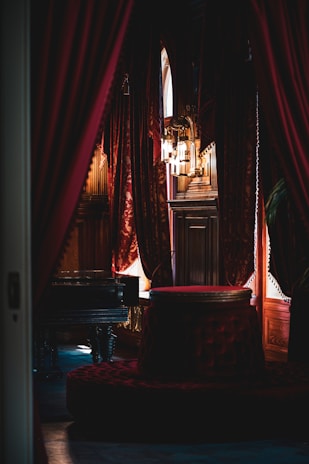 A dimly lit, opulent room with deep red velvet curtains and wooden paneling. A grand piano sits in the shadowy interior. The warm glow from multiple ornate light fixtures adds to the luxurious ambiance.