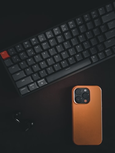 A mechanical keyboard with dark keys and one red key in the top left corner, arranged alongside a smartphone with a brown case. Both objects are set against a dark background, creating a minimalist and sleek appearance.
