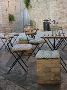 A rustic outdoor seating area featuring wooden chairs and stone-patterned cushions is arranged on a cobblestone street. The backdrop consists of stone walls with small windows and some greenery creeping over the edges. Lavender plants in metal pots are placed on tables, adding a touch of color to the scene.