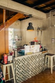 A cozy coffee shop counter with a geometric tile design. The countertop has various items such as a drink menu, a coffee machine, containers, and a 'self service' sign. The ambient lighting includes a pendant lamp with a warm glow, and there are wooden and concrete elements in the decor.