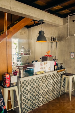 A cozy coffee shop counter with a geometric tile design. The countertop has various items such as a drink menu, a coffee machine, containers, and a 'self service' sign. The ambient lighting includes a pendant lamp with a warm glow, and there are wooden and concrete elements in the decor.