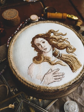 An intricately embroidered depiction of a woman with flowing hair on a white fabric background, framed in a circular wooden hoop. The image is surrounded by various vintage and decorative items, including spools of thread and patterned objects.