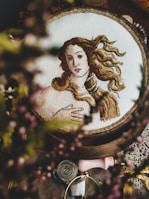 An embroidery hoop features a detailed cross-stitch depiction of a classical figure with flowing hair. The piece is surrounded by blurred elements that suggest an artistic workspace, including what appears to be lace, a magnifying glass, and small decorative items.