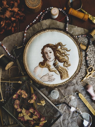 A detailed embroidery of a classical figure, resembling Renaissance art, is showcased in a wooden hoop. The scene includes various vintage sewing accessories and tools such as spools of thread, scissors, beads, and a magnifying glass. Dried flowers are neatly arranged in a glass frame, adding to the rustic aesthetic. The arrangement sits on a crinkled brown paper background, enhancing the nostalgic and artistic atmosphere.