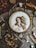 A detailed embroidery of a classical figure, resembling Renaissance art, is showcased in a wooden hoop. The scene includes various vintage sewing accessories and tools such as spools of thread, scissors, beads, and a magnifying glass. Dried flowers are neatly arranged in a glass frame, adding to the rustic aesthetic. The arrangement sits on a crinkled brown paper background, enhancing the nostalgic and artistic atmosphere.