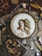 A detailed embroidery of a classical figure, resembling Renaissance art, is showcased in a wooden hoop. The scene includes various vintage sewing accessories and tools such as spools of thread, scissors, beads, and a magnifying glass. Dried flowers are neatly arranged in a glass frame, adding to the rustic aesthetic. The arrangement sits on a crinkled brown paper background, enhancing the nostalgic and artistic atmosphere.
