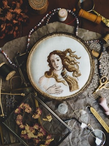 A detailed embroidery of a classical figure, resembling Renaissance art, is showcased in a wooden hoop. The scene includes various vintage sewing accessories and tools such as spools of thread, scissors, beads, and a magnifying glass. Dried flowers are neatly arranged in a glass frame, adding to the rustic aesthetic. The arrangement sits on a crinkled brown paper background, enhancing the nostalgic and artistic atmosphere.