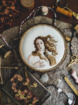 A detailed embroidery of a classical figure, resembling Renaissance art, is showcased in a wooden hoop. The scene includes various vintage sewing accessories and tools such as spools of thread, scissors, beads, and a magnifying glass. Dried flowers are neatly arranged in a glass frame, adding to the rustic aesthetic. The arrangement sits on a crinkled brown paper background, enhancing the nostalgic and artistic atmosphere.