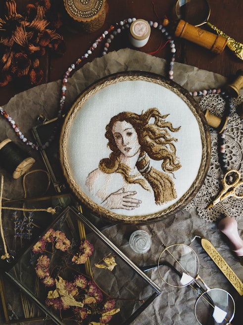 A detailed embroidery of a classical figure, resembling Renaissance art, is showcased in a wooden hoop. The scene includes various vintage sewing accessories and tools such as spools of thread, scissors, beads, and a magnifying glass. Dried flowers are neatly arranged in a glass frame, adding to the rustic aesthetic. The arrangement sits on a crinkled brown paper background, enhancing the nostalgic and artistic atmosphere.