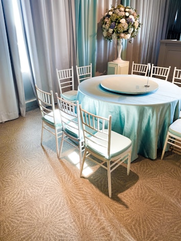 A round table covered with a light blue tablecloth, surrounded by elegant white chiavari chairs. A tall floral arrangement with mixed flowers, including white and blue, sits in a white vase nearby. The setting is indoors with soft natural light coming through tall curtains.