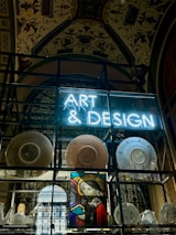 A display features a neon sign with the words 'ART & DESIGN' above shelves containing a variety of plates and pottery. A stained-glass artwork depicting a figure holding a book adds a historical and artistic touch. The background reveals an ornate ceiling adorned with elaborate patterns and designs.
