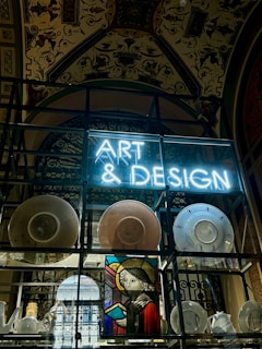 A display features a neon sign with the words 'ART & DESIGN' above shelves containing a variety of plates and pottery. A stained-glass artwork depicting a figure holding a book adds a historical and artistic touch. The background reveals an ornate ceiling adorned with elaborate patterns and designs.