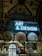 A display features a neon sign with the words 'ART & DESIGN' above shelves containing a variety of plates and pottery. A stained-glass artwork depicting a figure holding a book adds a historical and artistic touch. The background reveals an ornate ceiling adorned with elaborate patterns and designs.