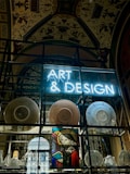 A display features a neon sign with the words 'ART & DESIGN' above shelves containing a variety of plates and pottery. A stained-glass artwork depicting a figure holding a book adds a historical and artistic touch. The background reveals an ornate ceiling adorned with elaborate patterns and designs.