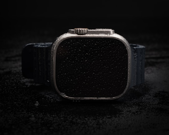 A black smartwatch with a sleek, rectangular face and a textured strap is covered in water droplets, set against a dark background, giving it a rugged and durable appearance.