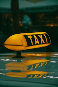 An illuminated taxi sign is mounted on the roof of a vehicle, reflecting brightly against a glossy surface. The letters are bold and clearly visible in a dark environment, suggesting nighttime or an area with low lighting.